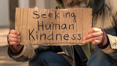Unhoused man holding sign that reads "Seeking human kindness"